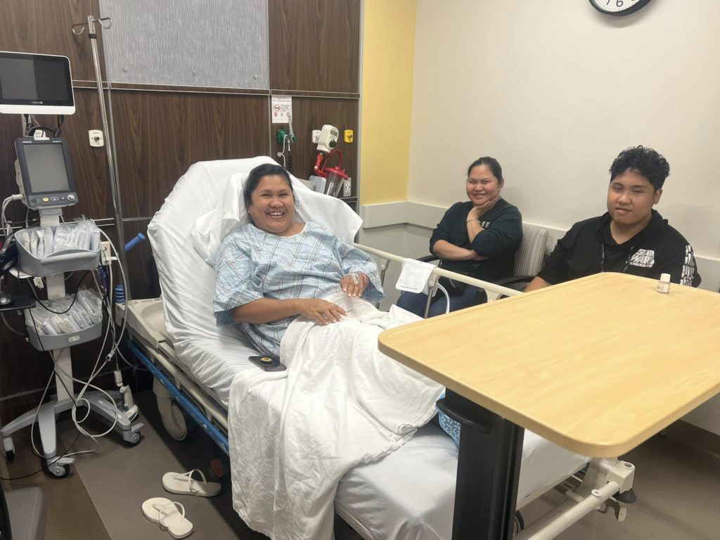 Patient smiling in a hospital bed with two visitors seated nearby in a bright room with medical equipment nearby