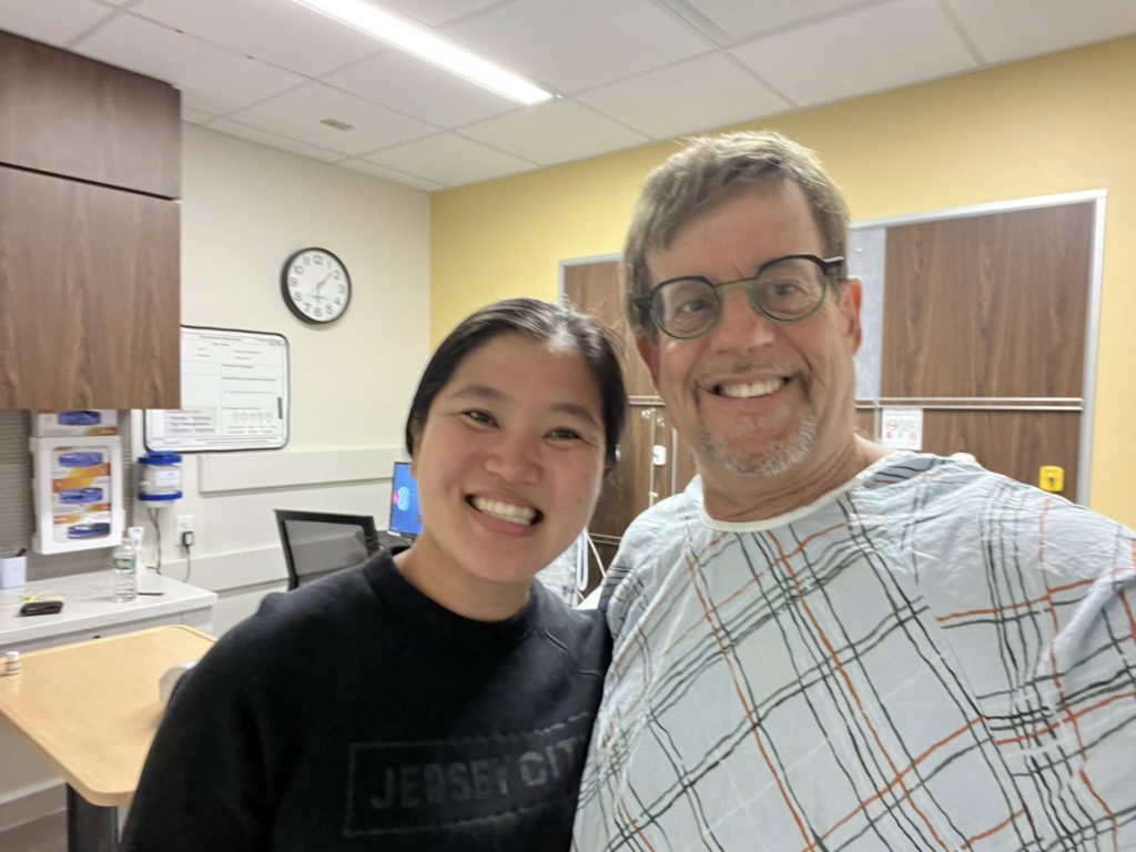 Two people smiling for a selfie in a hospital room, one in a black top and the other in a plaid hospital gown with orange and blue lines behind them.