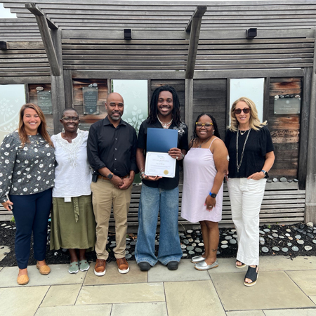 Tyrell-Weir_presentation | NJ Sharing Network Six people stand together outside a wooden building; central graduate holds a blue folder and certificate, flanked by friends and family.