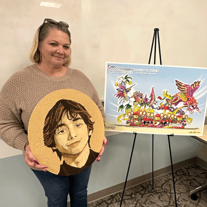 Woman in a beige sweater holds a round wood artwork featuring a portrait of a boy; an easel beside her displays a colorful poster.