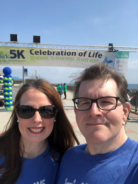 James-and-Tara-Condon-at-NJSN-5K | NJ Sharing Network Two people in blue shirts pose for a selfie on a sunny boardwalk under a banner reading '5K Celebration of Life' with the ocean in the background.