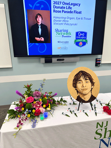 Table with a flower bouquet and a circular portrait cutout beside a projection screen about the 2027 OneLegacy Donate Life Rose Parade Float.