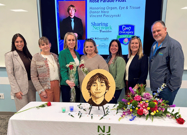 Eight people stand behind a memorial table with a circular portrait of a young man, flowers, and a blue tribute screen in the background honoring an organ donor hero.