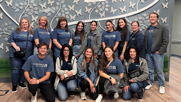 WIL---Clyde-Paul-Insurance-Agency-1 | NJ Sharing Network Group of colleagues wearing matching blue shirts posing for a team photo in front of a decorative butterfly mural in an office space.